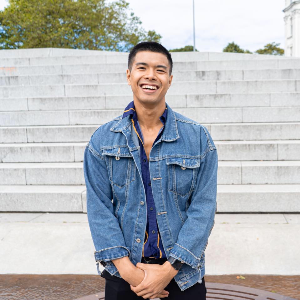 Graduate Artit Chatthai smiling on outdoor steps in a denim jacket